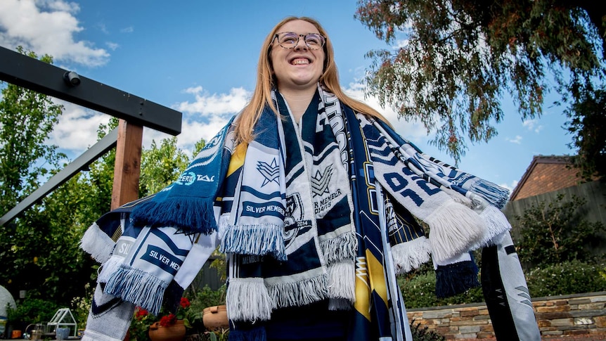 A young woman stands with several Melbourne Victory scarves around her neck.