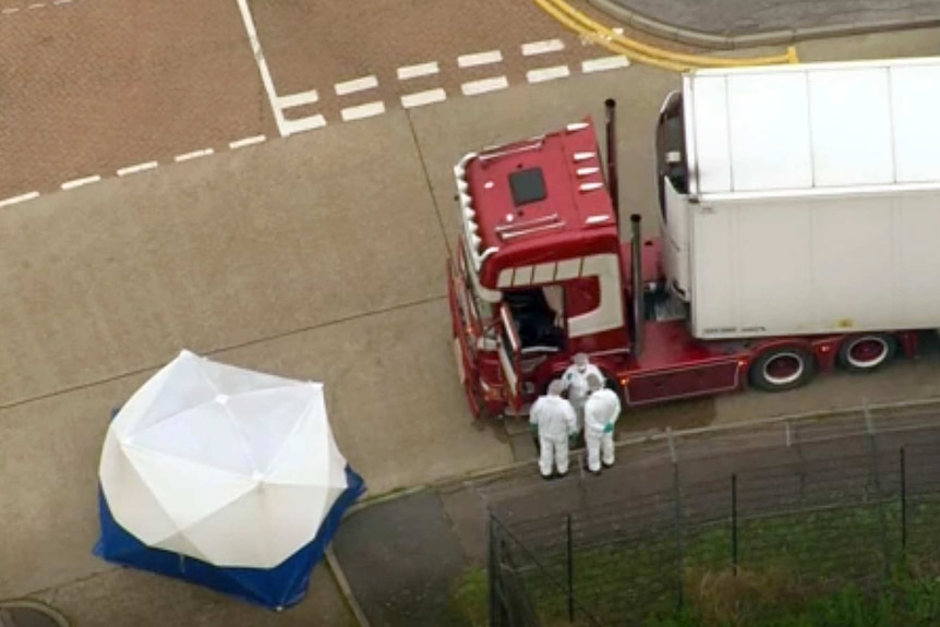 Forensic workers are seen from above standing next to a red truck with a white body. A tent has been erected to the left.