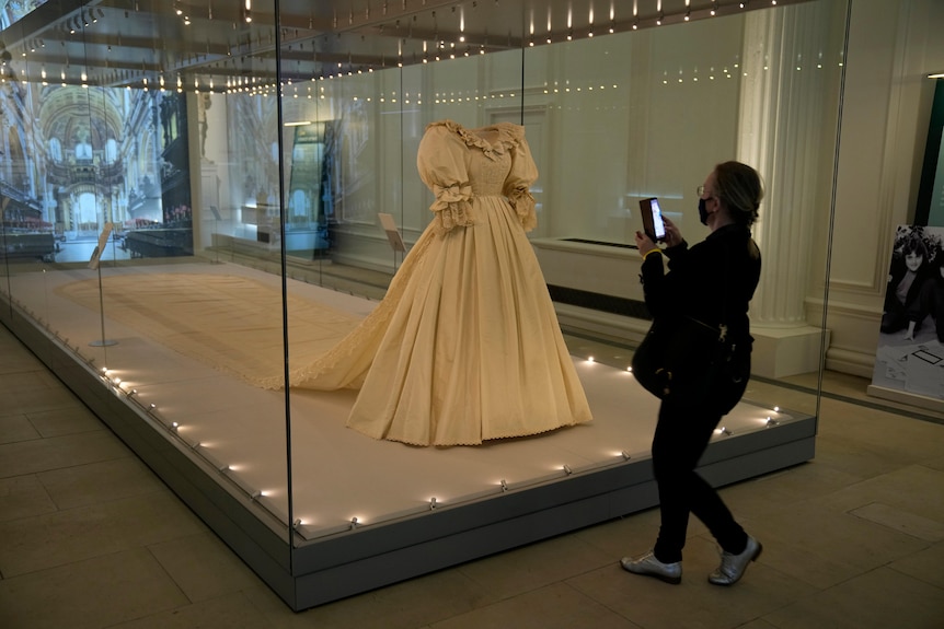 A woman takes a photo of Princess Diana's wedding dress, which is in a glass display case