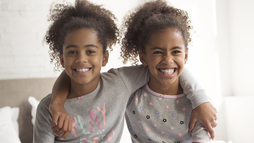 Two smiling young Black girls sitting on a bed in their pyjamas with their arms around each other’s shoulders.