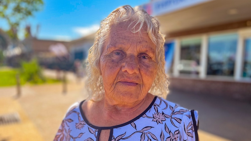 Elderly Indigenous woman with white hair and a flowery shirt looks into the camera, while standing on a street in front of a sh