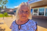 Elderly Indigenous woman with white hair and a flowery shirt looks into the camera, while standing on a street in front of a sh