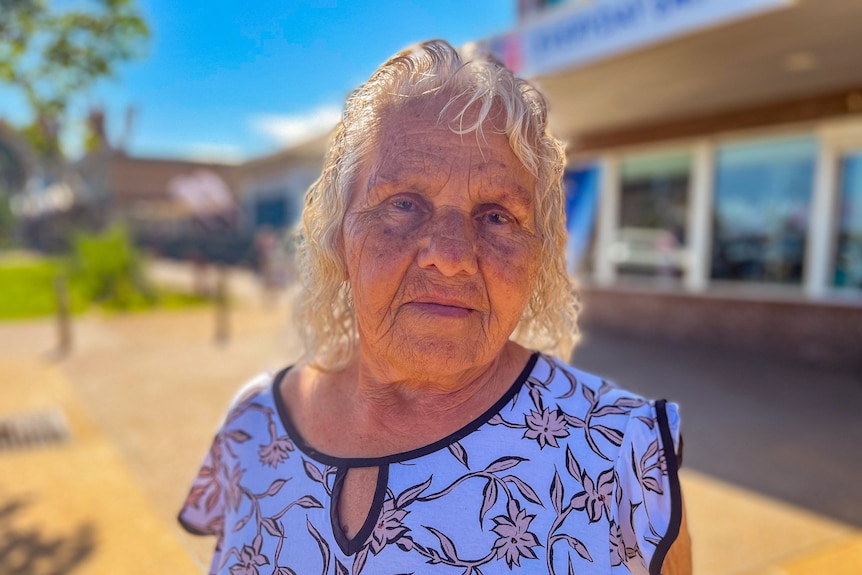 Elderly Indigenous woman with white hair and a flowery shirt looks into the camera, while standing on a street in front of a sh