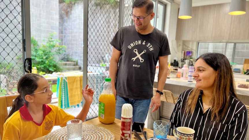 A woman and her daughter wearing her yellow school uniform sit at their table, the dad stands nearby.