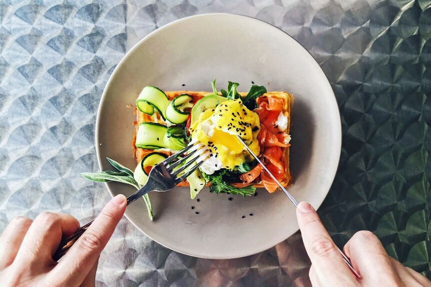 Hands holding cutlery hover over a white plate, cutting open a poached egg. Salmon and thin zucchini slices sit beside the egg.