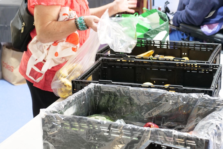 Crates of capsicums and bananas sit next to each other on a table, with a woman filling a plastic bag with bananas to take.