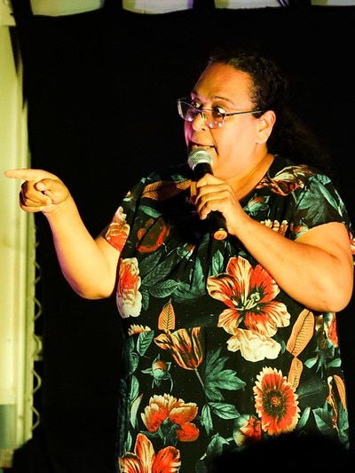 Indigenous female comedian on stand-up comedy stage. She has a microphone in her hand and her arm is out-stretched