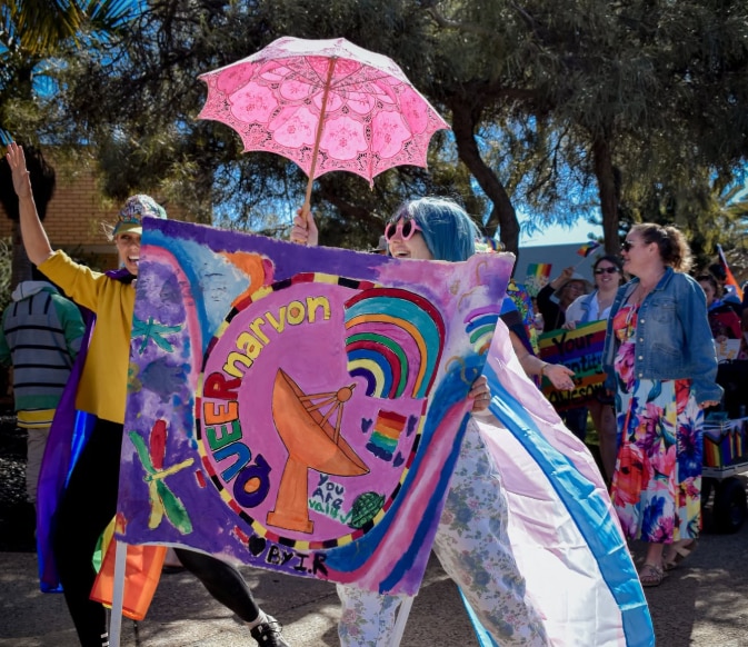 A woman in a blue wig with a sign reading: Queernarvon.