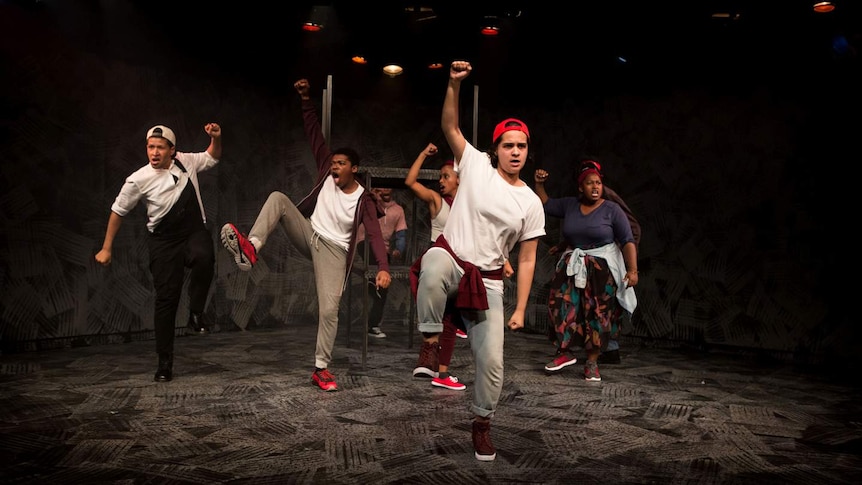 Seven performers on stage dressed in casual clothes shout with a raised fist.