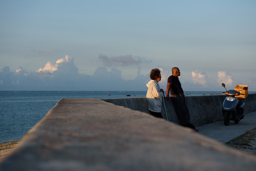 an elderly couple stand by a stone fence on the shoreline in Japan