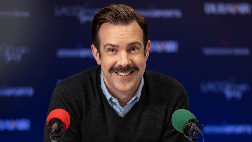 A 40-something man with a moustache smiles while seated at a press conference in front of one red and one green microphone.