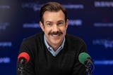 A 40-something man with a moustache smiles while seated at a press conference in front of one red and one green microphone.