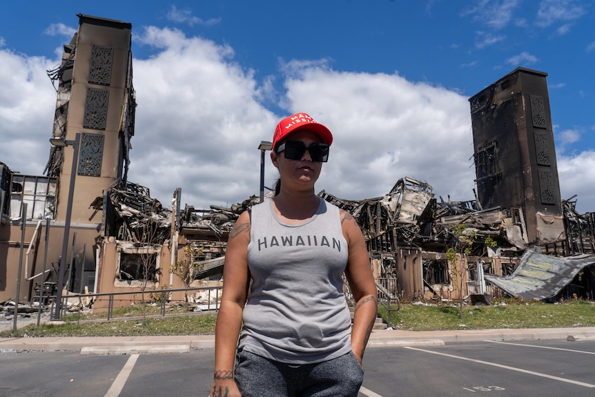 Tiffany, wearing a red cap, sunglasses, and a grey tank top that says 'Hawaiian', stands in front of a destroyed building.