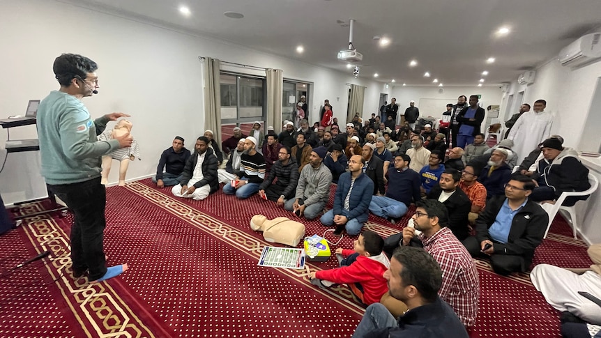 A man gives a lesson to people seated on the floor of a mosque