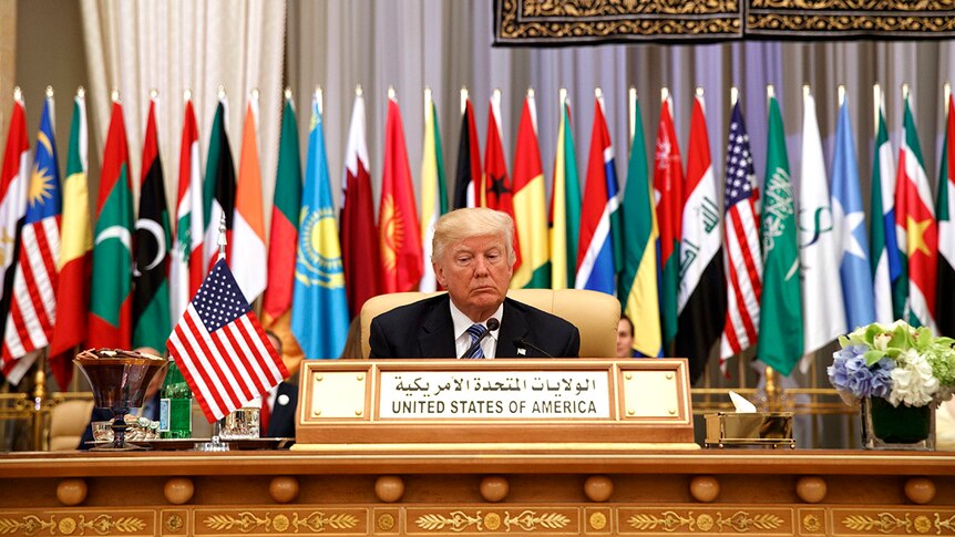 Donald Trump sits at a table, behind him are flags of Muslim nations