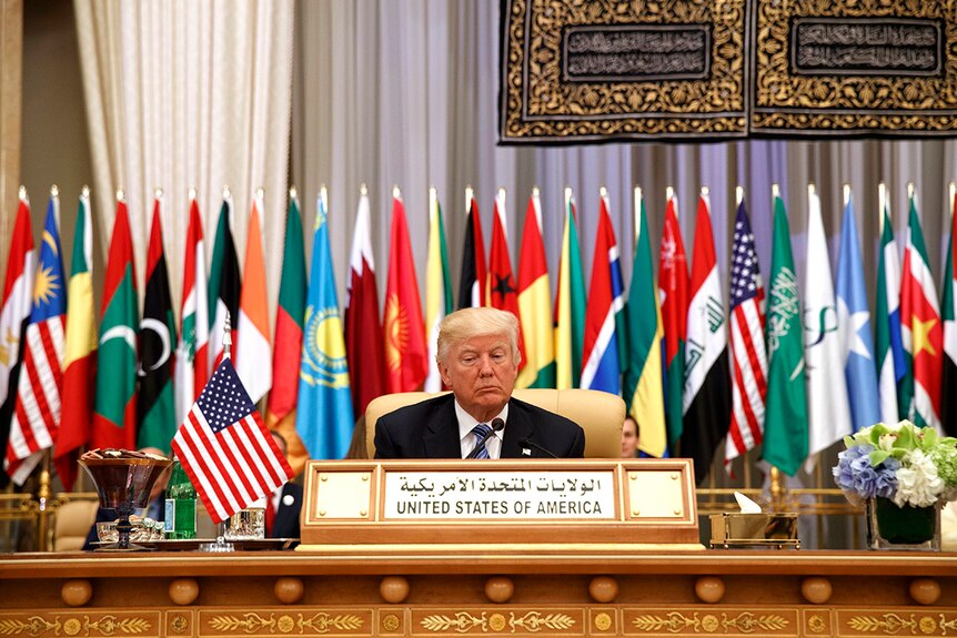 Donald Trump sits at a table, behind him are flags of Muslim nations