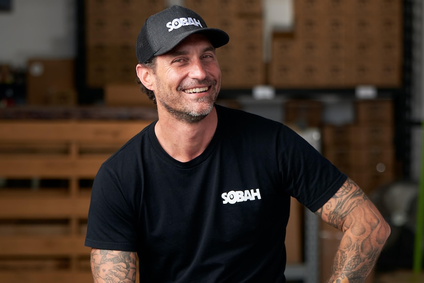 A man wearing a black shirt and cap and jeans sitting on a bar stool.