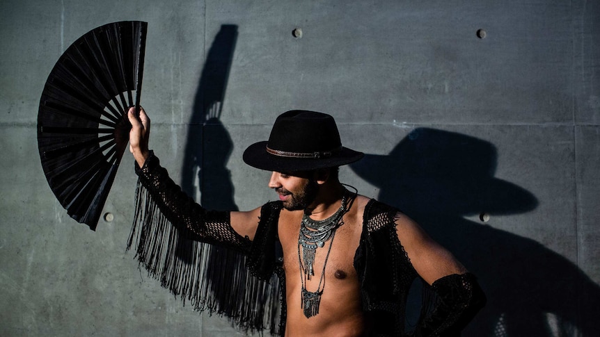 Man in black hat, holding a black fan, in front of a grey wall.