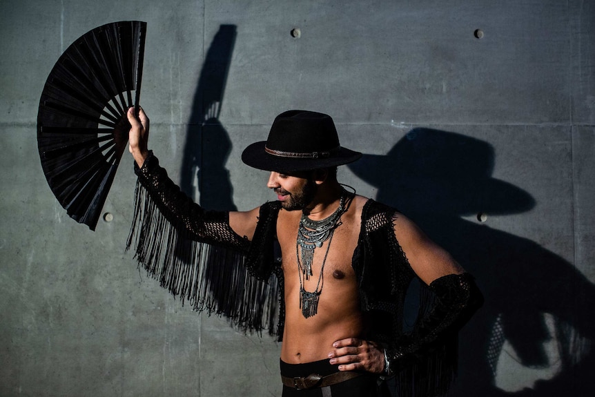 Man in black hat, holding a black fan, in front of a grey wall.