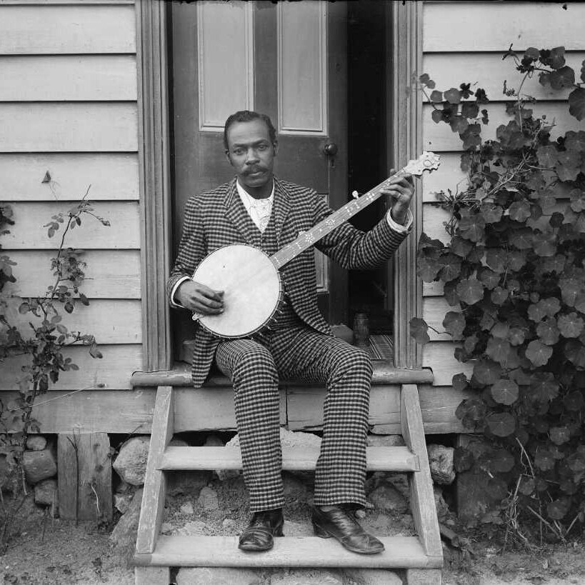 A black man sits on the front steps of a wooden house playing the banjo wearing a checked suit