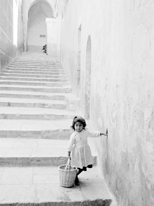 A black and white photograph shows a small girl with a basket as stone steps climb behind her.