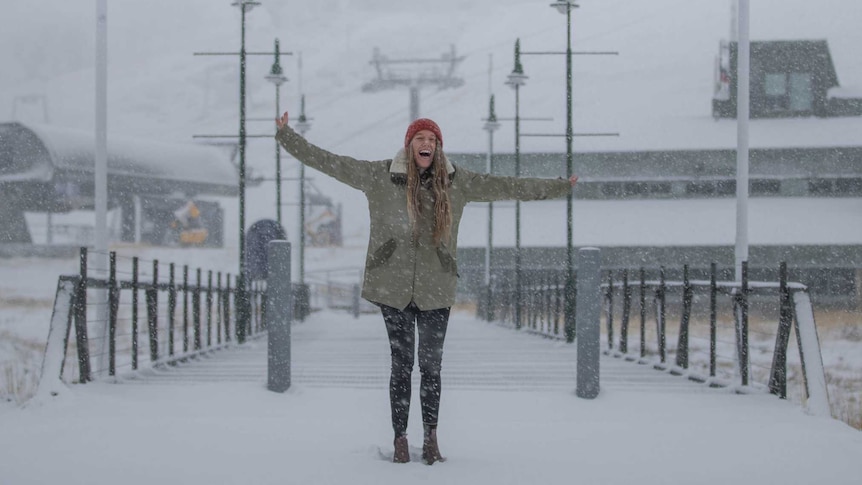 A laughing woman stands with her arms wide open on a bridge as snow falls around her.