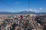 A red cable car passes over a favela in Rio de Janeiro, a blue sky and mountains are in the background