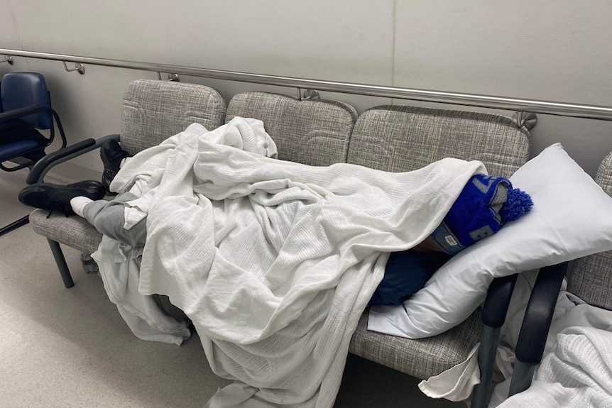 A teenager wearing a beanie, hoodie and track pants, sits against a wall in a hospital  while charging his mobile phone