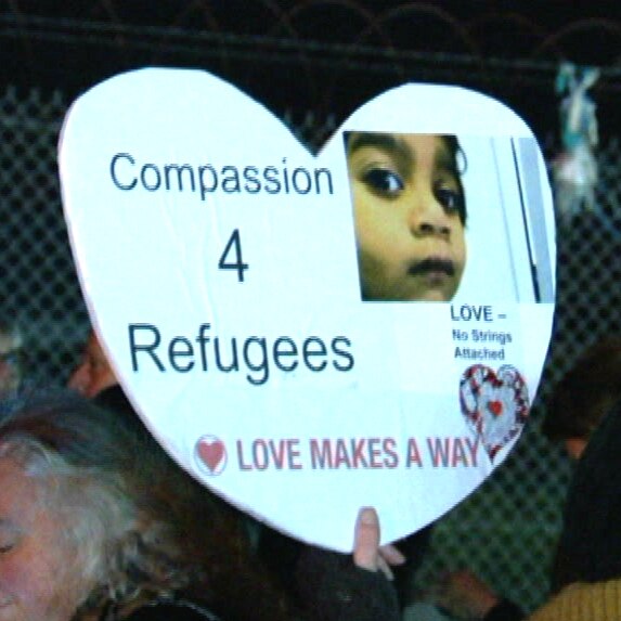 A white sign in the shape of a love heart bears the words 'Compassion 4 Refugees' with an image of a child.