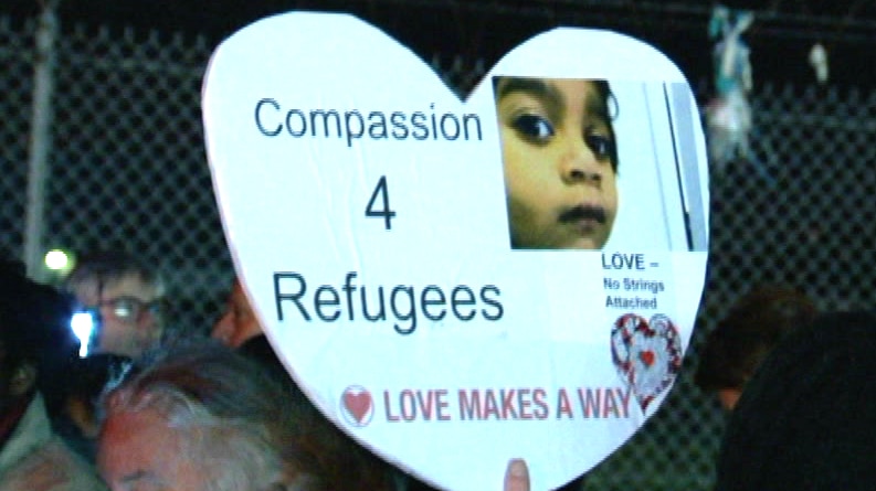 A white sign in the shape of a love heart bears the words 'Compassion 4 Refugees' with an image of a child.