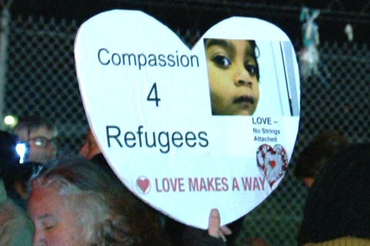 A white sign in the shape of a love heart bears the words 'Compassion 4 Refugees' with an image of a child.