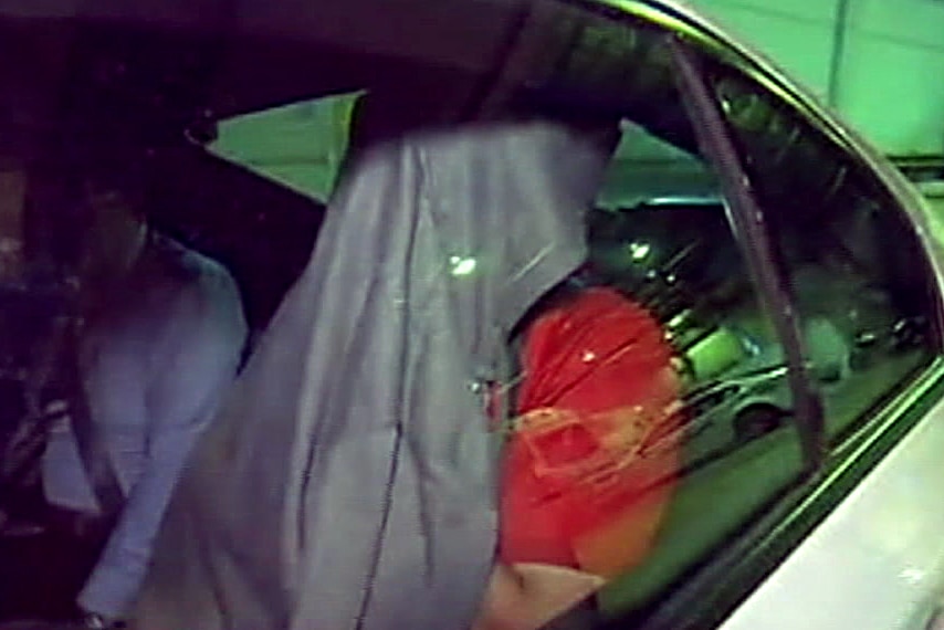 A man in the back of a police car with a red t-shirt and a grey jacket over his head.