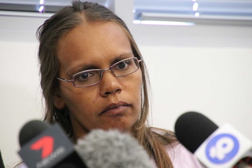 A woman wearing glasses at a press conference in front of a bank of microphones.
