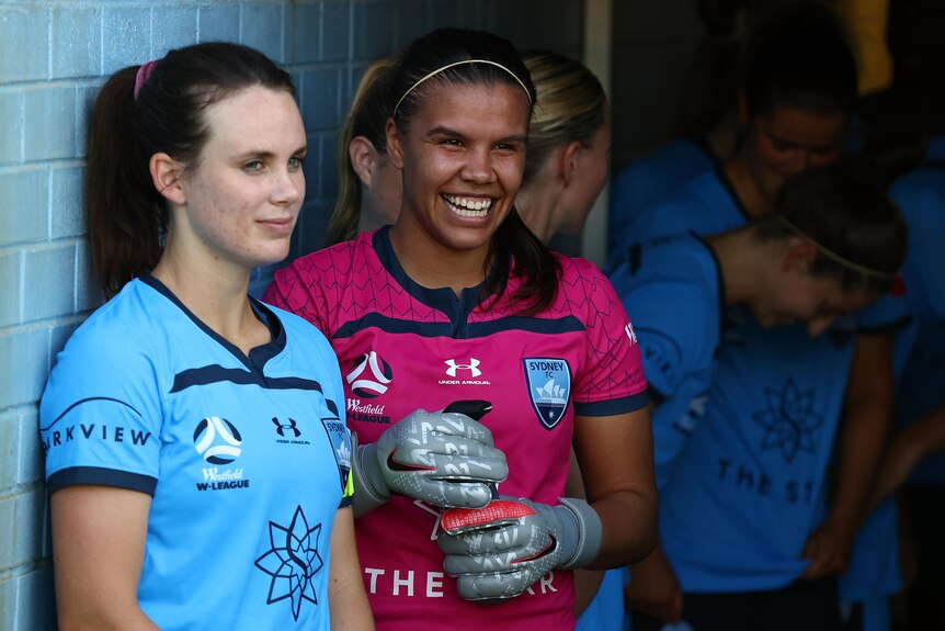 Women soccer players wearing blue and pink stand against a wall smiling