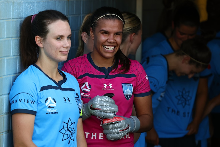 Women soccer players wearing blue and pink stand against a wall smiling