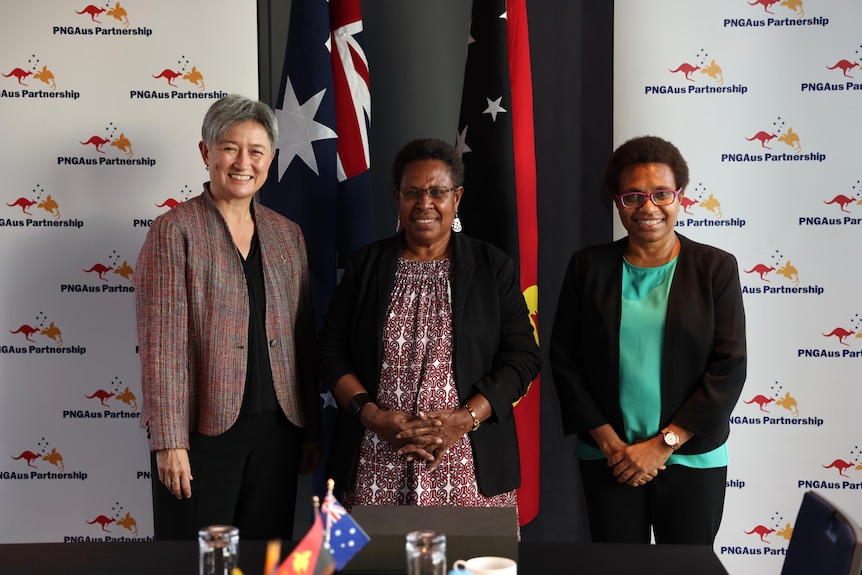 Three women, including Penny Wong, stand side by side in front of an Australian and Papua New Guinea flag