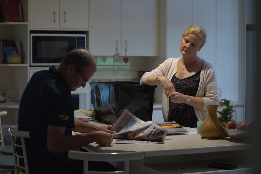 A man sits and a woman stands in a kitchen looking down at documents on the bench