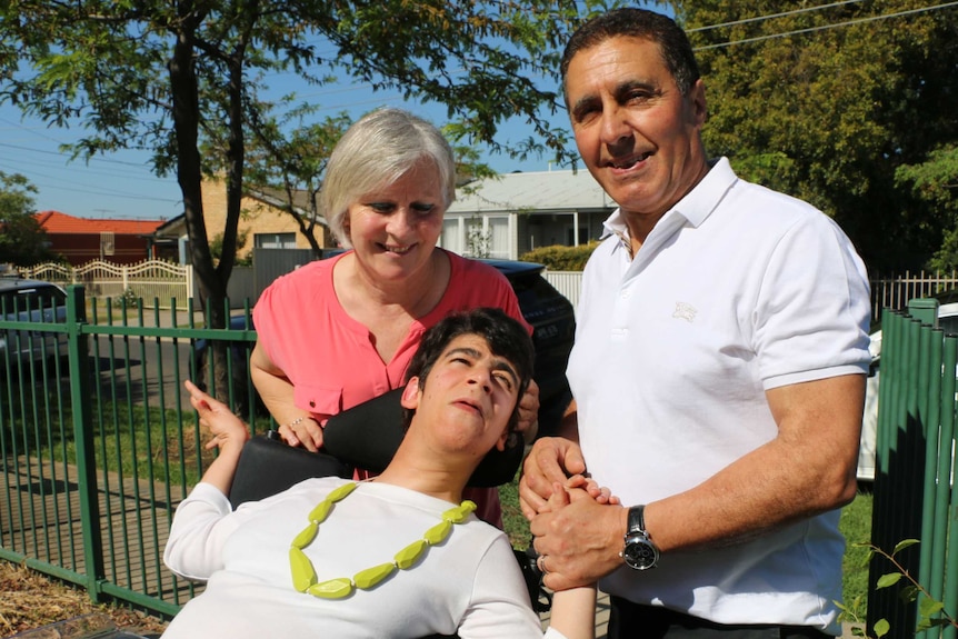 Liana Indovino, sits in her wheelchair, reaching up to hold the hands of her parents.