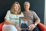 Older couple sit on lounge holding framed picture of son