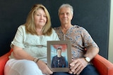 Older couple sit on lounge holding framed picture of son