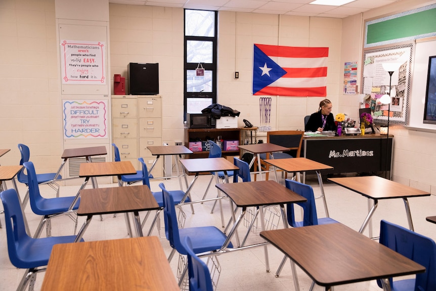 A teacher sits at a desk in a classroom with rows of empty desks. 