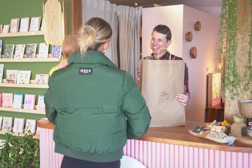 A woman with short hair hands a paper bag across a counter to a woman in a green puffer jacket.