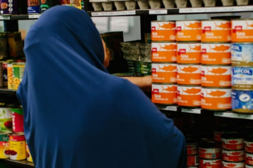 A photo showing cans on a shelf, and a woman wearing a dark blue hijab facing the shelf. 