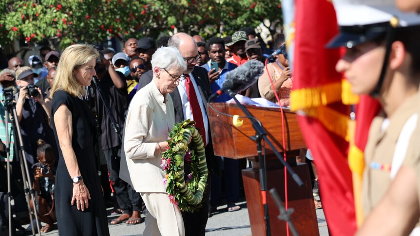 A woman holds a wreath, flanked by another man and woman, with a soldier to the right of the image.