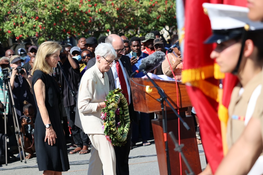 A woman holds a wreath, flanked by another man and woman, with a soldier to the right of the image.