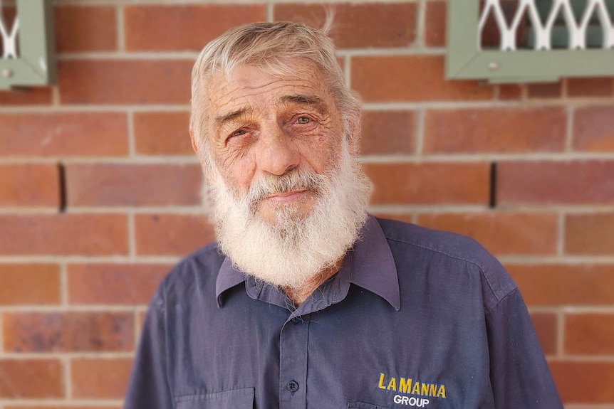 A man with white hair and a beard, wearing a navy shirt, standing against a brown brick wall.