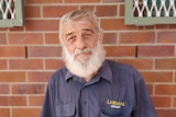 A man with white hair and a beard, wearing a navy shirt, standing against a brown brick wall.