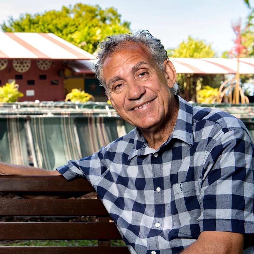 An Indigenous man with curly grey hair, wearing a check shirt, sitting on a park bench, with a building in the background.