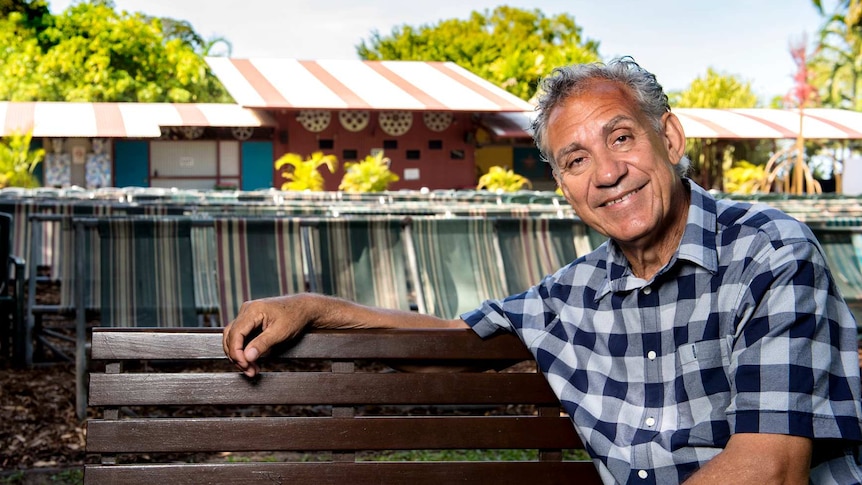 An Indigenous man with curly grey hair, wearing a check shirt, sitting on a park bench, with a building in the background.