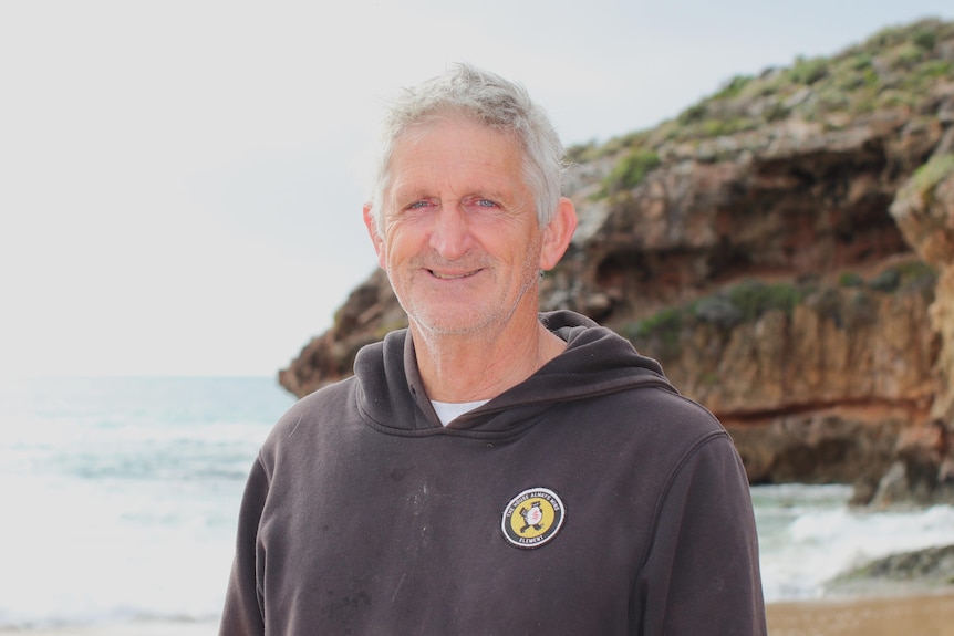 A grey-haired smiling man with ocean and cliff in background. Wears a black hoodie with yellow logo.